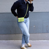 27 of 27:A person standing outdoors against a gray concrete wall, wearing a dark navy jacket, light blue jeans, tan sneakers, and carrying a bright yellow-green envelope-style crossbody bag with a thin yellow strap.