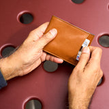 12 of 20:A close-up of a person's hands holding a slim, tan leather bifold wallet with a card partially inserted into its side slot, set against a dark red textured background with circular cutouts.