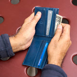 14 of 20:A person's hands holding open a dark blue leather bifold wallet, showing card slots, a credit card, and a dollar bill, set against a red perforated metal background.