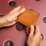 13 of 20:A close-up of a person's hands holding a slim, tan leather card holder wallet against a textured reddish-brown background with circular cutouts.