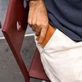 16 of 20:A close-up of a person's hand sliding a tan leather cardholder or wallet into the pocket of off-white trousers, while seated on a dark red chair.