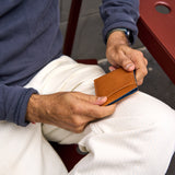 17 of 20:A close-up of a man holding a slim, tan leather bifold wallet with a blue fabric lining or insert, seated on a white chair.