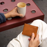 18 of 20:A person holding a slim tan leather bifold wallet in one hand while sitting at a red metal table and holding a cup with coffee in the other hand.