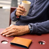 19 of 20:A tan leather bifold wallet with a blue interior lining resting on a reddish-brown metal table, with a person in a navy shirt holding a cup in the background.