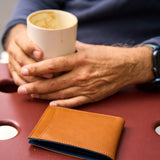 20 of 20:A tan leather bifold wallet with a blue interior lining resting on a reddish-brown table, with a person's hands holding a white ceramic mug in the background.
