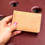 2 of 6:A tan snakeskin-textured leather card holder or wallet held by a hand with pink nail polish against a dark reddish-brown textured background.