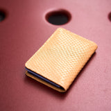 4 of 6:A slim, tan textured card holder wallet with a python-skin style pattern, holding several cards, resting on a dark red textured surface with circular cutouts.