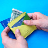 15 of 23:A close-up of a person's hands holding a lime green leather card holder wallet, showing it filled with cards and a dollar bill, against a solid blue background.