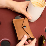 2 of 8:A person holds a slim brown leather wallet with a snap-button flap, shown in a real-life setting with a coffee cup and sunglasses on a dark red table.