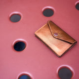 8 of 12:A small metallic bronze leather wallet with a flap closure and snap button, resting on a textured pink surface with circular cutouts.