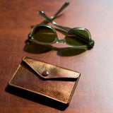 9 of 12:A pair of green-tinted sunglasses resting on a dark wooden table next to a small brown leather card holder with a snap button closure.