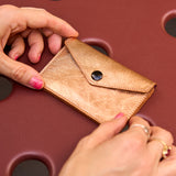 2 of 12:A close-up of a small tan leather card holder or wallet with a flap closure and snap button, being held by two hands over a dark reddish-brown textured surface.