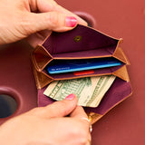 3 of 12:A close-up of a tan leather wallet being opened by two hands, showing a deep purple interior with card slots holding cards and a dollar bill.