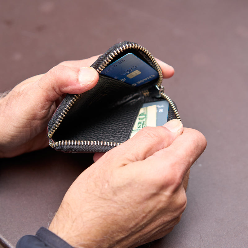 Person opening a black leather wallet with money and cards on a brown surface