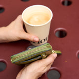 8 of 39:A person holds a small olive green zip-around wallet in one hand and a paper coffee cup in the other, set against a dark red outdoor table with circular cutouts.