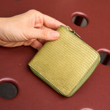 34 of 39:A hand holding a small zip-around wallet with a textured olive green exterior and gold-toned zipper hardware, set against a dark red plastic surface with circular cutouts.