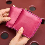 29 of 39:A close-up of a person's hands holding a small, vibrant magenta leather zip-around wallet with gold-tone hardware, set against a dark reddish-brown textured background with circular cutouts.