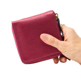 10 of 39:A hand holding a small textured leather zip-around wallet in a deep magenta pink color, shown against a plain white background.
