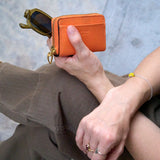 3 of 9:A person holding an orange leather card holder wallet with gold-tone hardware and sunglasses, wearing brown pants and a silver bracelet against a gray concrete background.