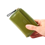 26 of 34:An olive green leather TUSK zip-around wallet being held by a hand against a plain white background.