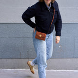 4 of 27:A person walking outdoors against a gray concrete wall, wearing a dark navy jacket, light blue jeans, tan shoes, and carrying a brown leather crossbody bag with a gold snap closure.
