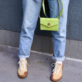 26 of 27:A person wearing light blue jeans and tan suede sneakers with white laces, carrying a bright lime green envelope-style crossbody bag with a gold-tone turn-lock closure, standing against a textured gray wall.