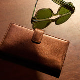 2 of 15:Green-tinted sunglasses with a matching brown leather case on a warm, reflective brown surface.