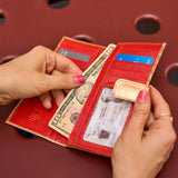 12 of 15:An red interior of a  bifold wallet with gold trim, shown open with a hand inserting a ten-dollar bill and another hand holding an ID card. The wallet contains multiple card slots, a cash pocket, and a gold-tone snap closure.