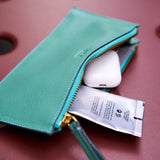 3 of 10:A teal leather zip pouch with gold-tone hardware, shown open with a white earbud case and a small tube of cream inside, set against a dusty rose background.