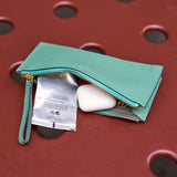 2 of 10:A mint green leather zip pouch with a wrist strap, shown open with a silver packet and a white earbud case inside. The pouch is placed on a textured red surface with circular cutouts.