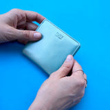 2 of 14:A light blue leather bifold wallet with the TUSK logo embossed on the front, held by two hands against a solid bright blue background.
