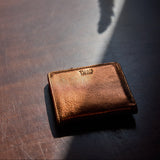 9 of 17:A small brown leather wallet with a debossed 'TUGS' logo, resting on a dark wooden surface under dramatic, direct sunlight that casts a strong shadow.