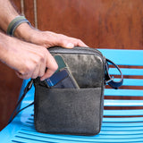 3 of 10:Person holding a black leather crossbody bag on a blue chair with a wooden background