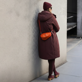 7 of 23:Person wearing a long brown coat and red beanie leaning against a wall.