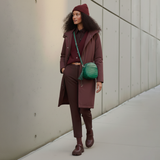 2 of 16:Woman in a maroon outfit with a green bag standing against a neutral wall.