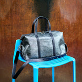 2 of 12:A dark gray distressed leather duffel bag with black leather handles and trim, resting on a bright blue plastic chair against a textured rusted metal wall.