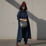 2 of 13:Woman wearing a long denim coat, maroon beanie, and sunglasses against a plain wall.