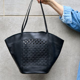 4 of 7:Black leather tote bag with woven pattern held by a person against a gray wall.