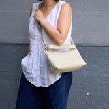 3 of 8:Person wearing a white sleeveless top and blue jeans, holding a beige handbag against a gray wall.