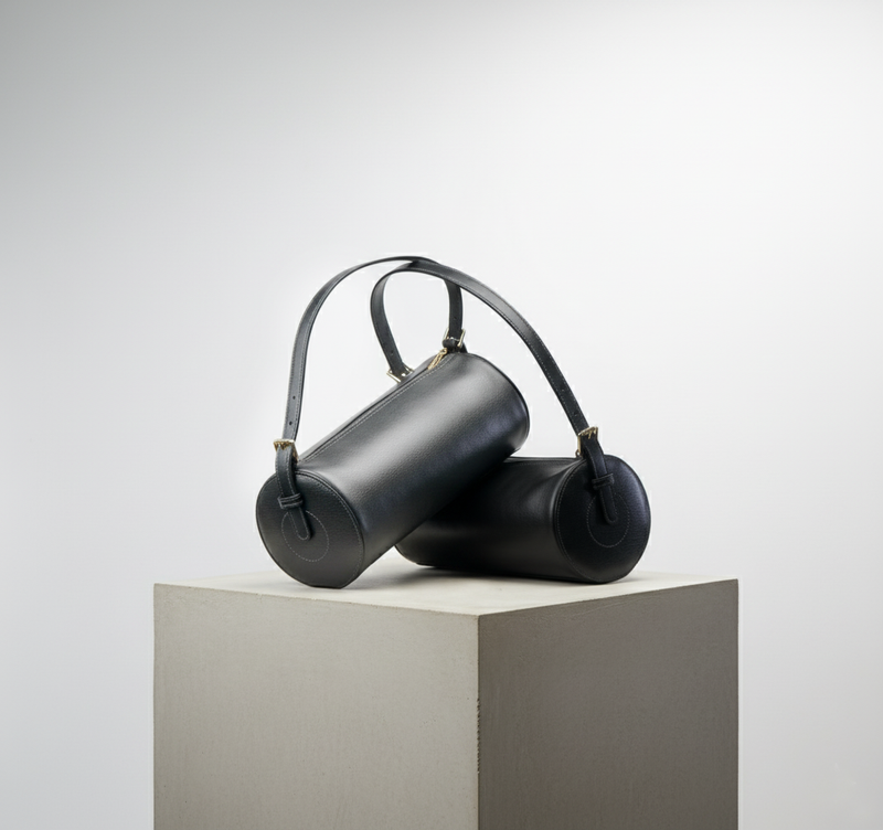 Black cylindrical handbag on a white pedestal against a light gray background