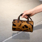 3 of 43:A hand holding a small cylindrical handbag with a brown and black leopard print pattern and dark brown handles, set against a concrete pavement background - CAMEL/BLACK