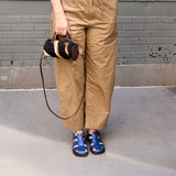 16 of 43:A person wearing tan wide-leg pants and blue sandals holds a small black, brown, and beige striped cylindrical shoulder bag with a long strap, standing against a gray brick wall.
