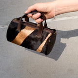 18 of 43:A hand holding a small cylindrical handbag with a black and tan zebra-patterned hair-on-hide body and dark brown leather handles, set against a gray asphalt background.