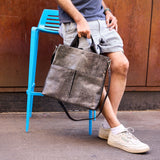 10 of 16:Person sitting on a blue stool holding a gray leather bag against a brown wall.