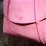 5 of 20:A close-up of a pink suede TUSK small shoulder bag with a flap closure and visible 'TUSK NEW YORK' branding, set against a dark textured background -  BLUSH