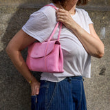 6 of 20:A woman wearing a white t-shirt and blue pinstripe pants carries a pink suede shoulder bag against a textured gray concrete wall -  BLUSH
