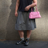 8 of 20:A person wearing a dark gray pleated midi skirt, black t-shirt, gray socks, and black Salomon trail running shoes, carrying a pink leather shoulder bag against a textured concrete wall -  BLUSH
